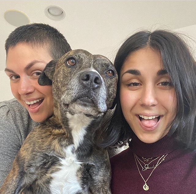 Yasmine Yousaf in a maroon high neck sweater smiling with her pet dog.